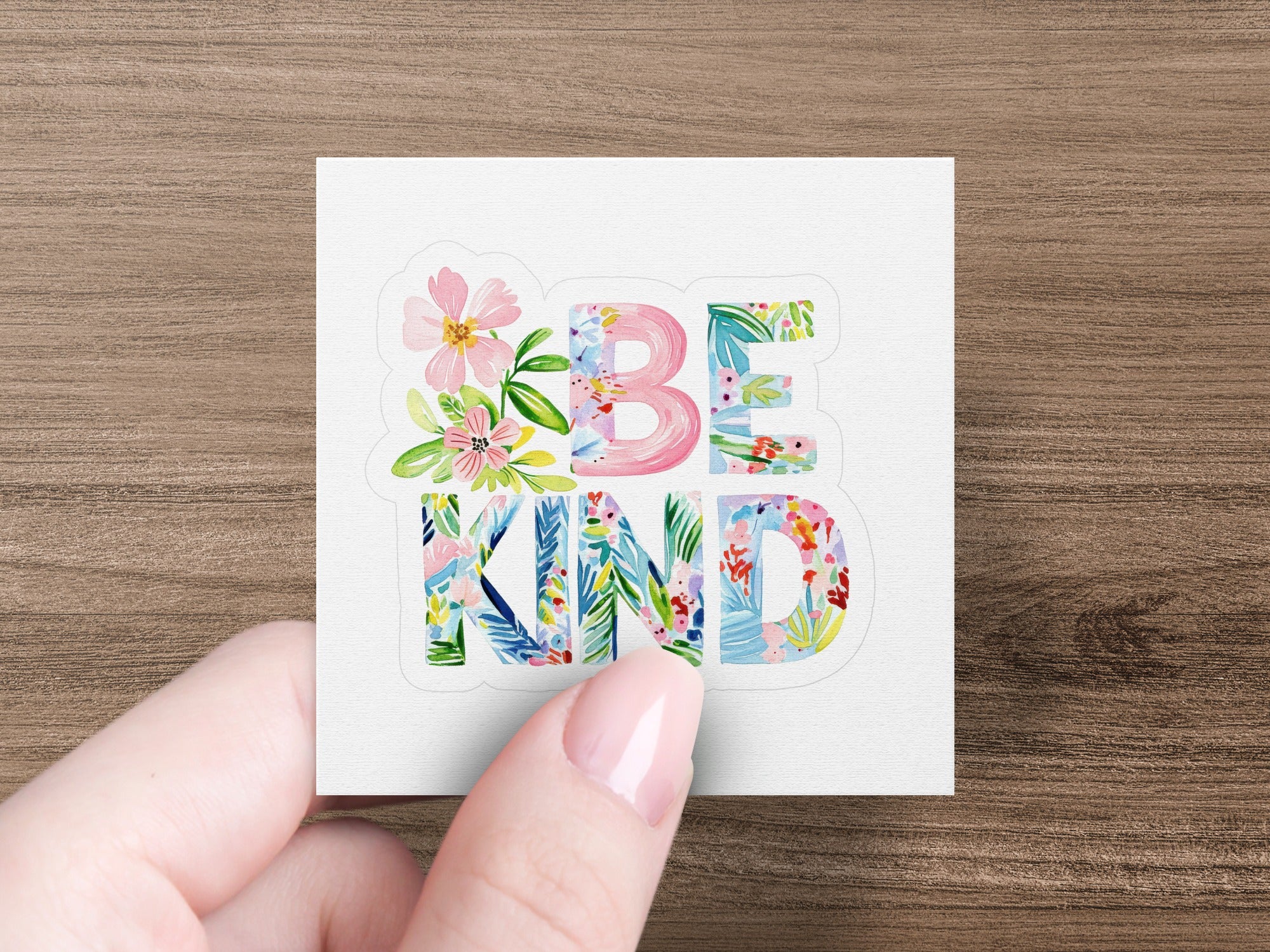 Floral "BE KIND" text on a white square sticker held by a hand.