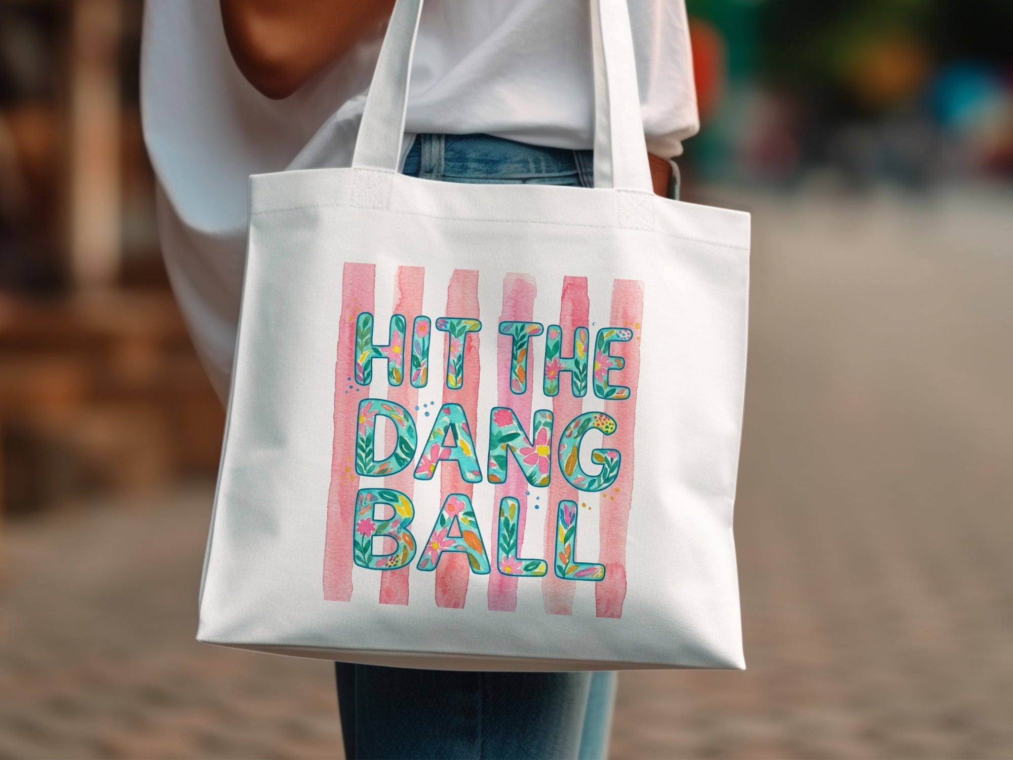 White tote bag with pink stripes and floral text reading "HIT THE DANG BALL".