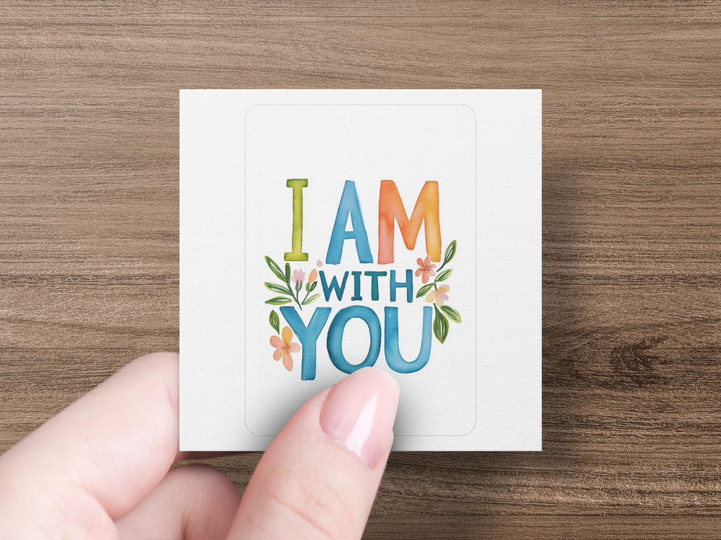 I AM WITH YOU text with floral accents on a white square card held by a hand.