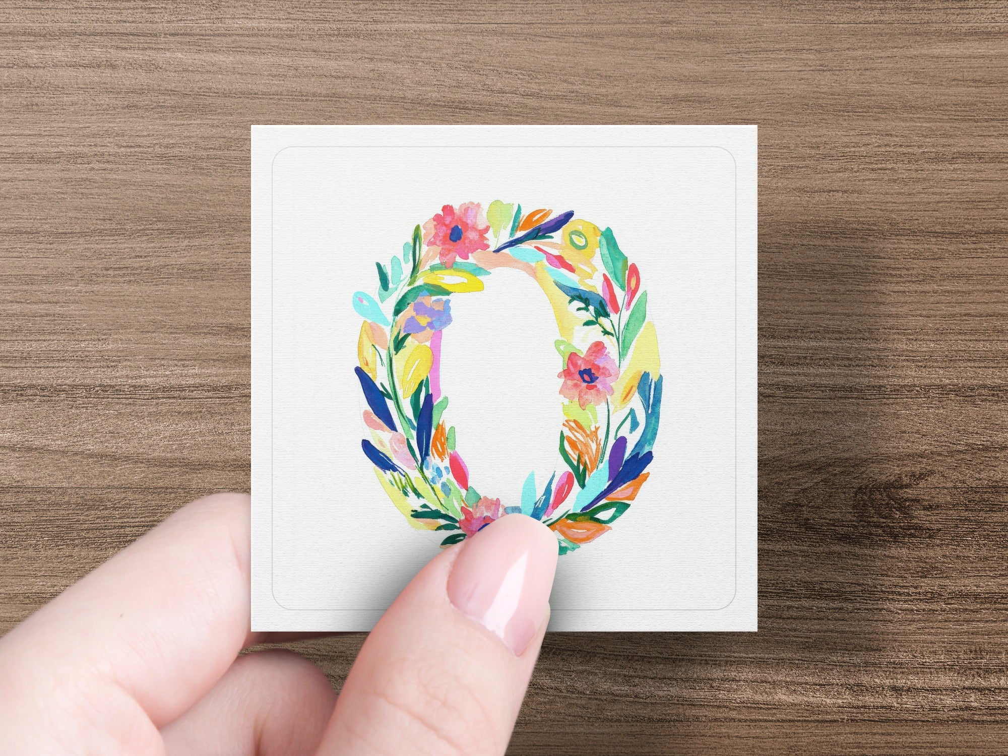 Hand holding square card with colorful floral letter O illustration.