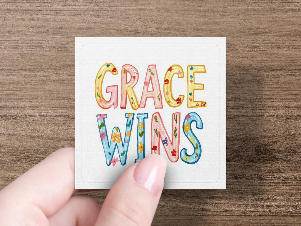 "GRACE WINS" text in colorful, floral lettering on a square card held by a hand.