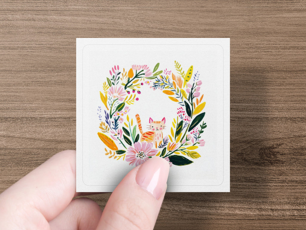 Square card with floral wreath and smiling cat illustration held by a hand.