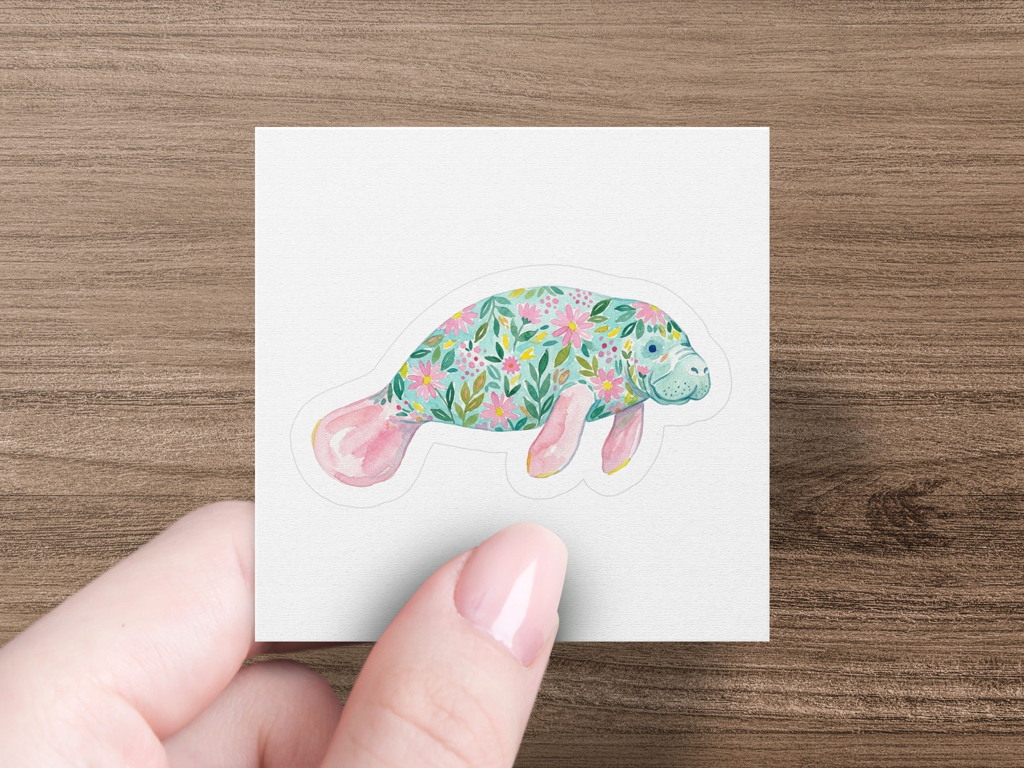 Manatee sticker with floral pattern held by fingers on a wooden background.