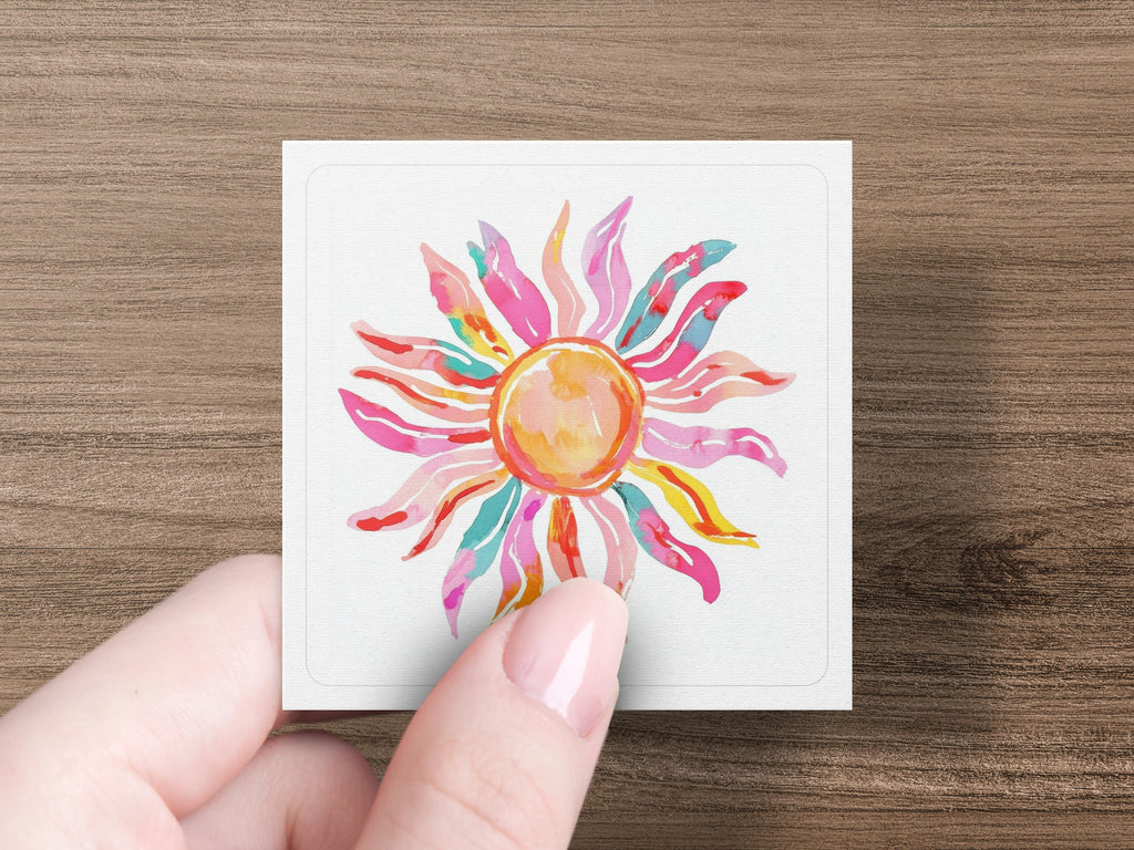 Pink and yellow watercolor sun on a white square held by a hand.