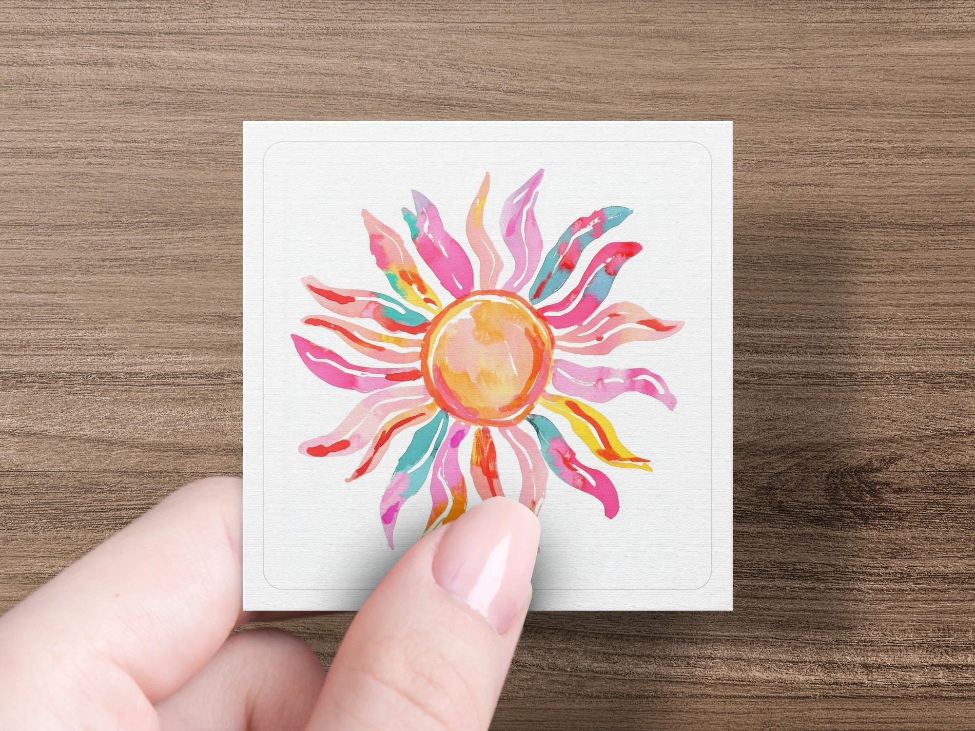 Pink and yellow watercolor sun on a white square held by a hand.