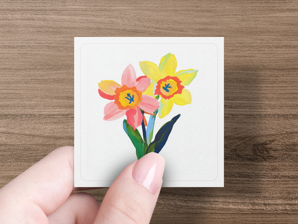 Two watercolor daffodils, one pink and one yellow, held by a hand.