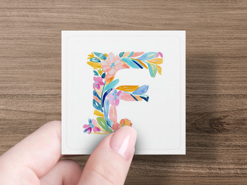 Watercolor floral letter 'F' on a white card held by a hand.