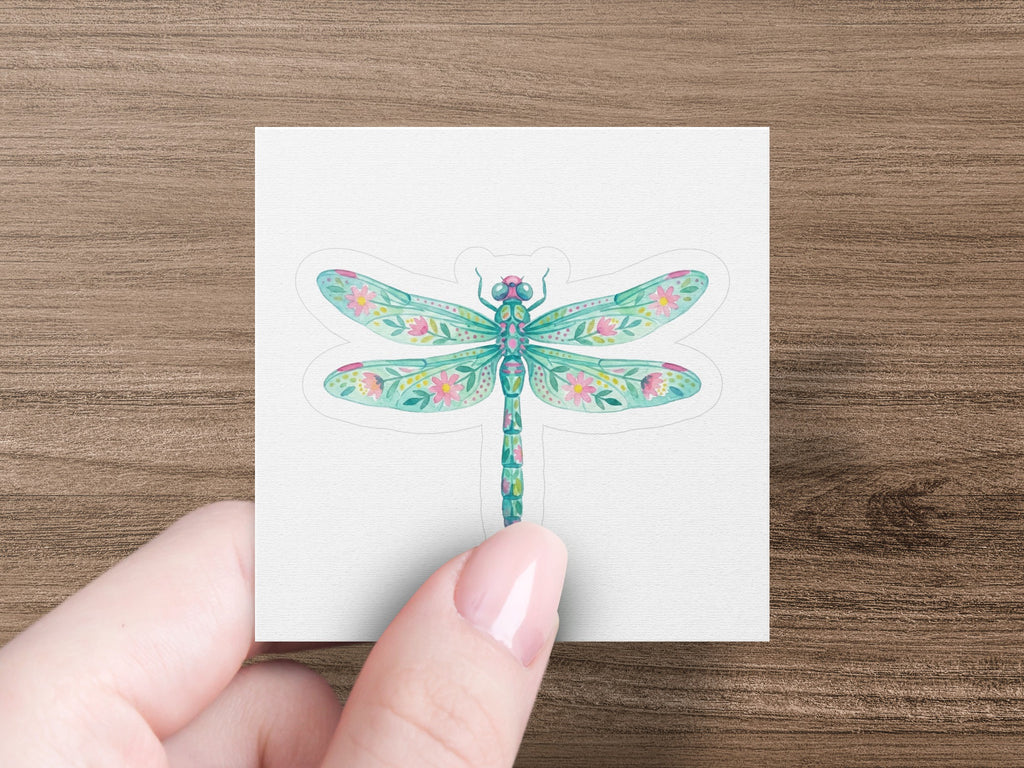 Dragonfly sticker with floral wing patterns on white paper held by a hand.
