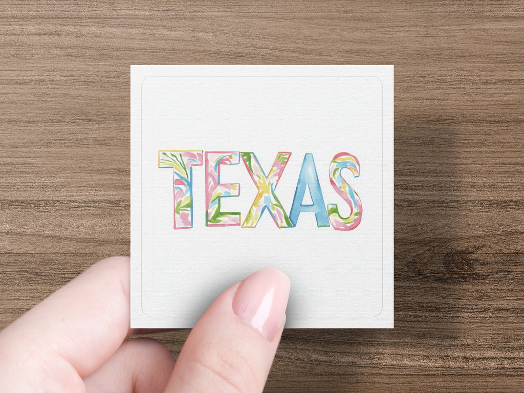 Watercolor letters spell "TEXAS" on a square card held by a person's fingers.