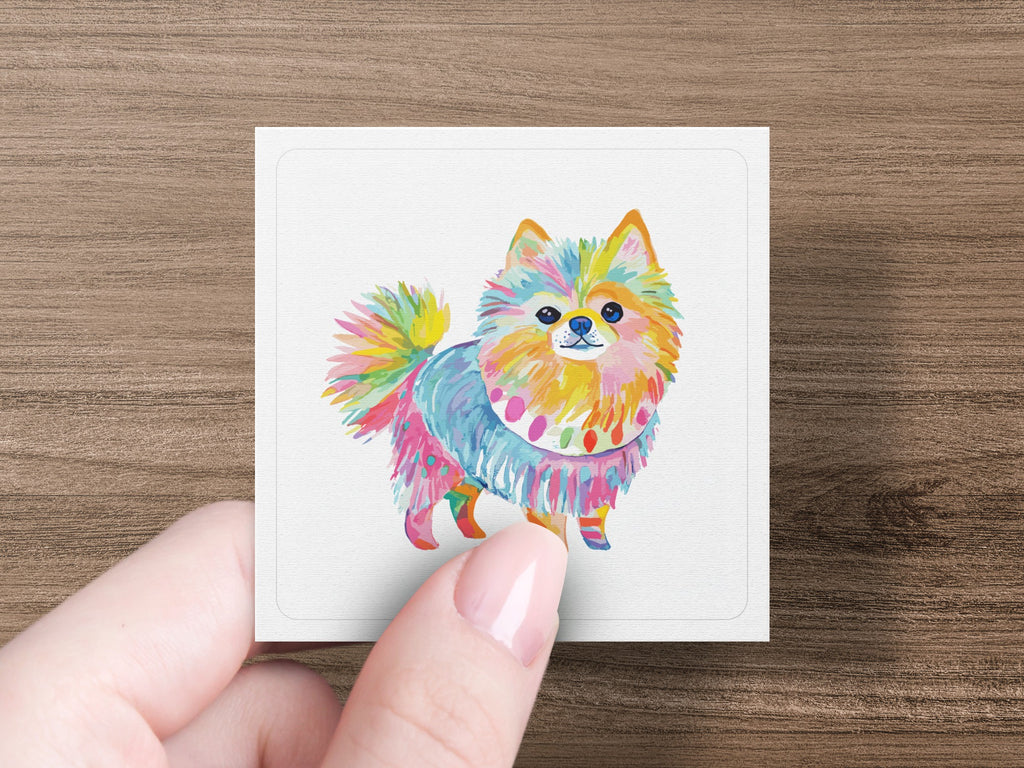Hand holding colorful, painted Pomeranian dog artwork on white card.
