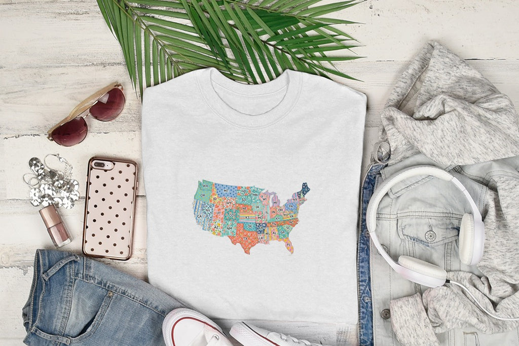 White t-shirt with colorful US states map design, surrounded by headphones and clothing.