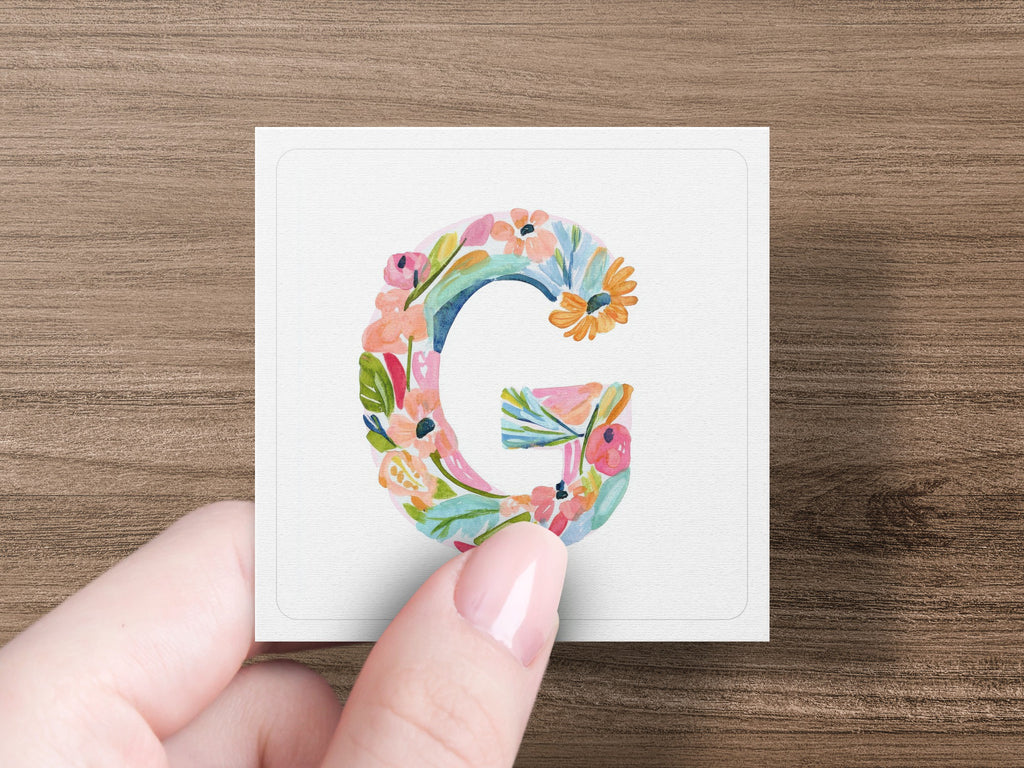 Hand holding square card with floral letter 'G' illustration.