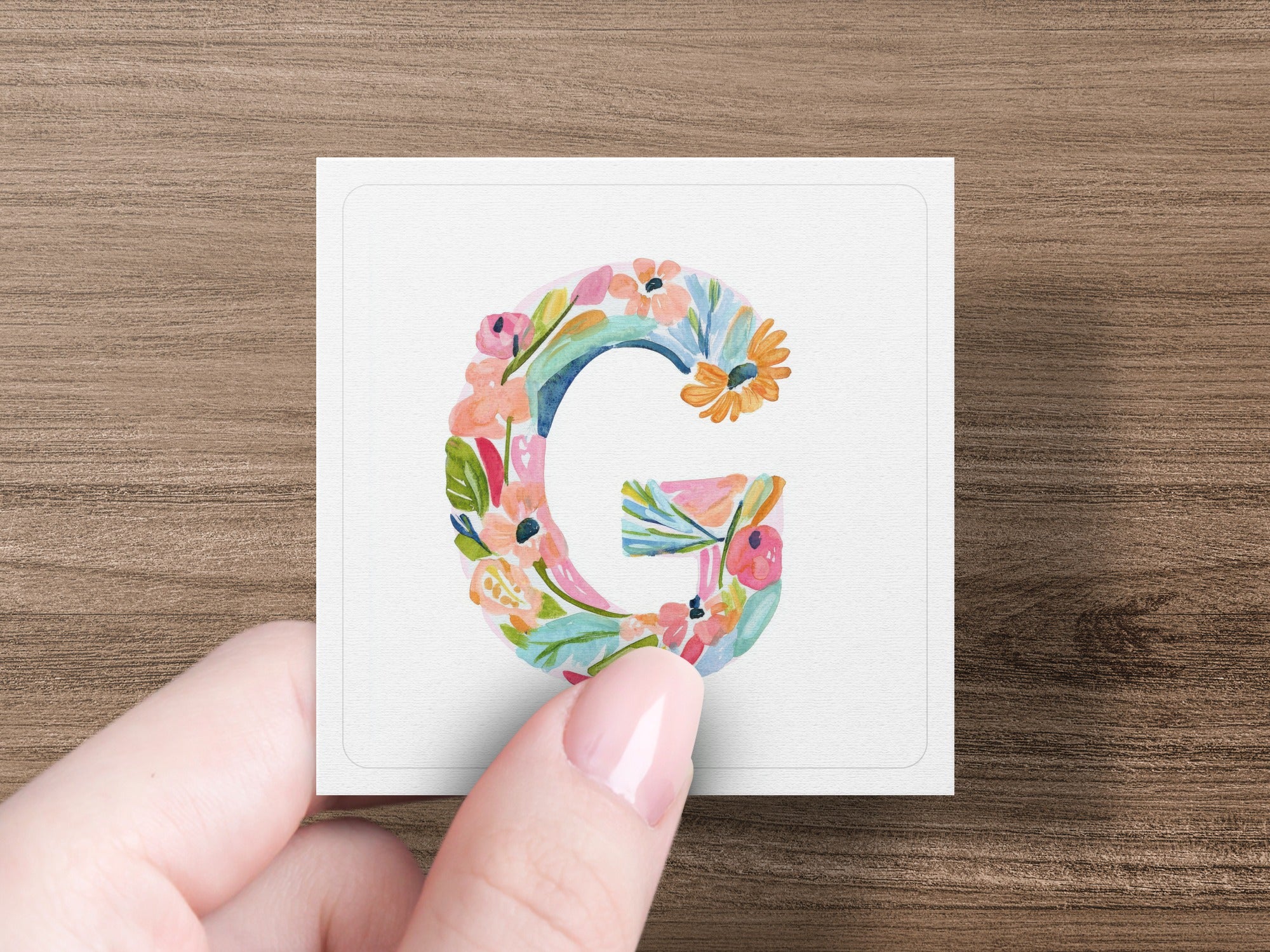 Hand holding square card with floral letter 'G' illustration.