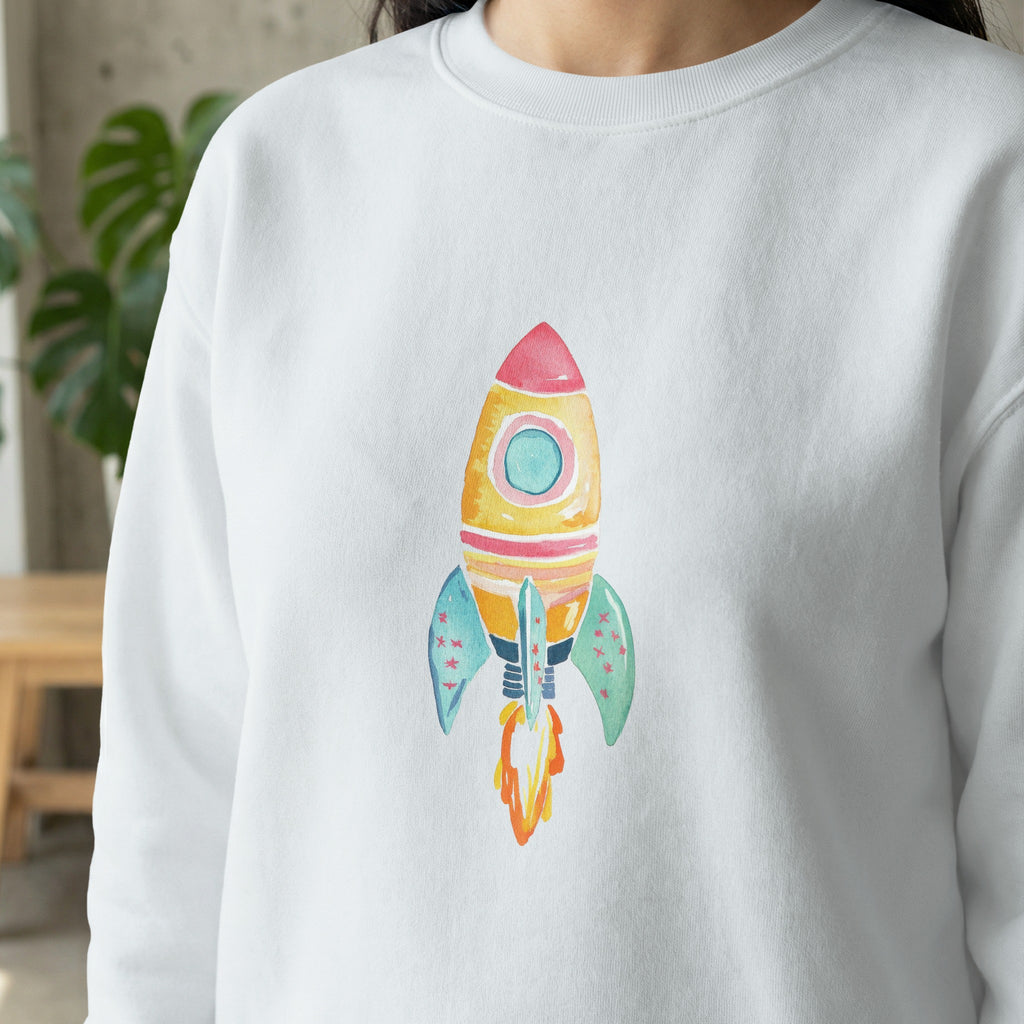 White sweatshirt featuring a watercolor illustration of a colorful rocket ship.