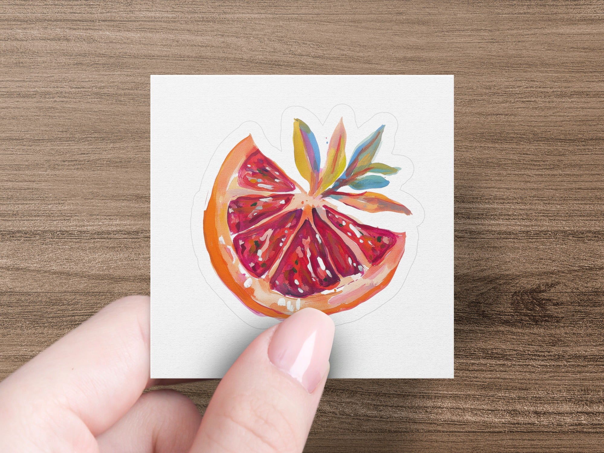 Hand holding a sticker of a sliced, ripe grapefruit with colorful leaves.