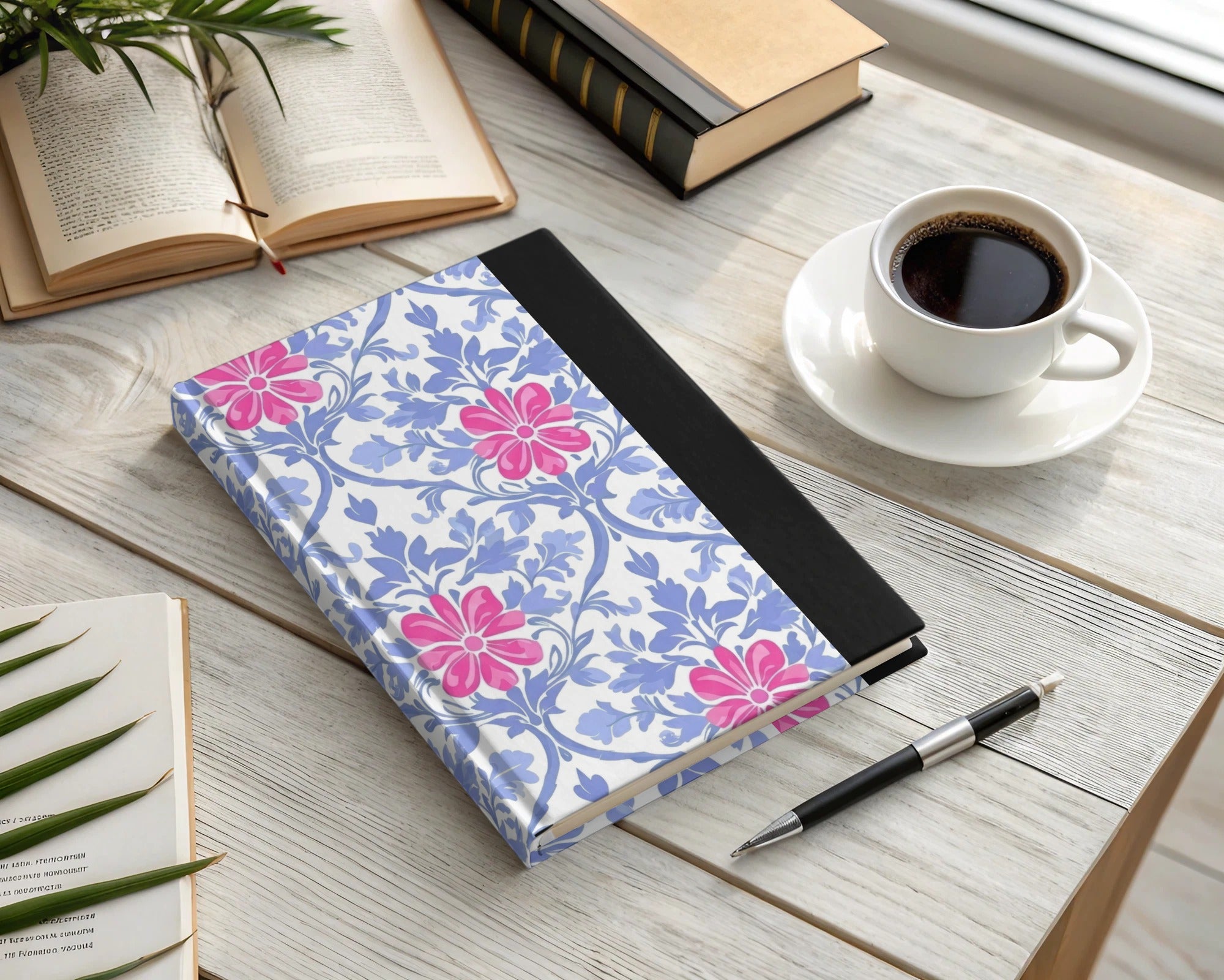 Floral notebook with pink flowers and blue leaves on a desk with a pen and coffee.