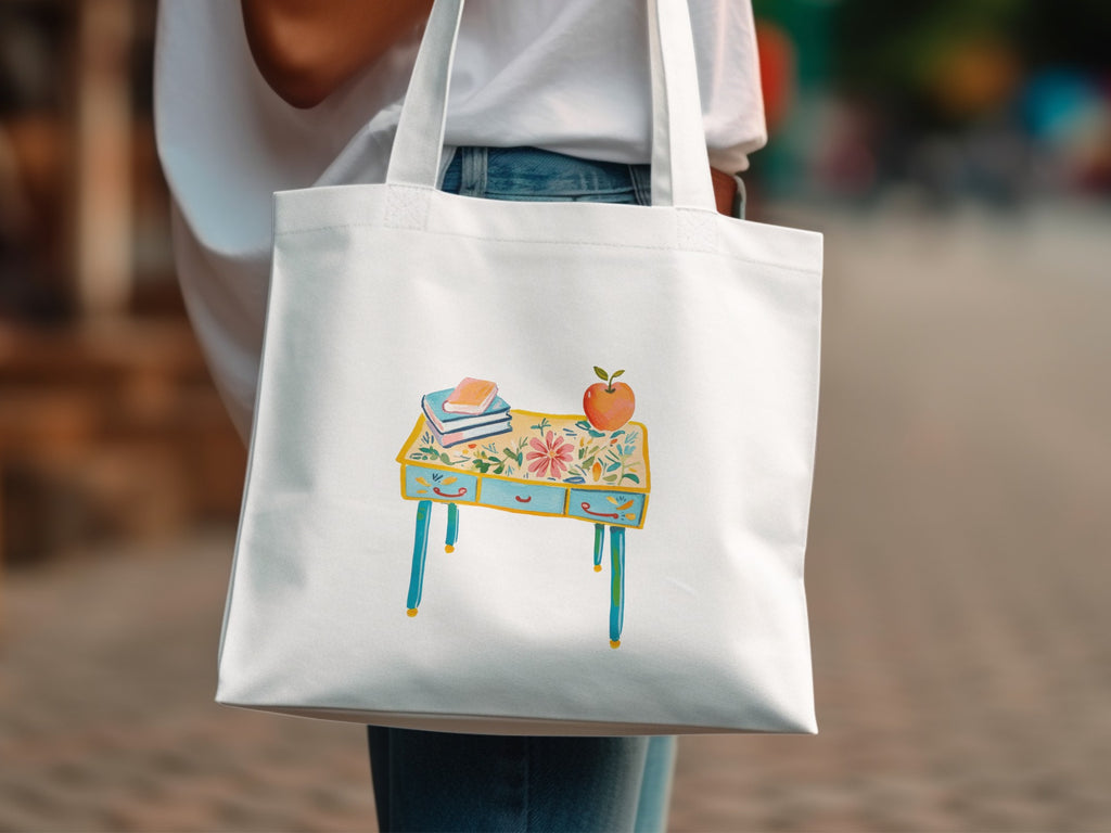 White tote bag with painted desk, books, and apple design.