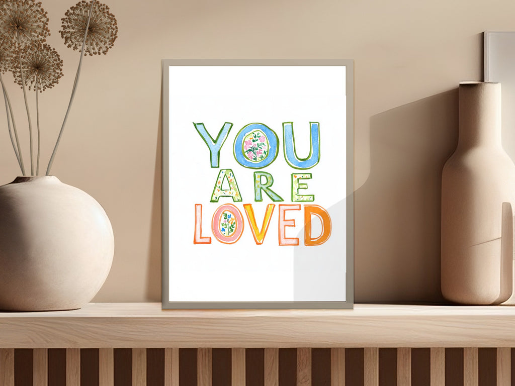 Framed art with colorful "YOU ARE LOVED" text on a shelf.