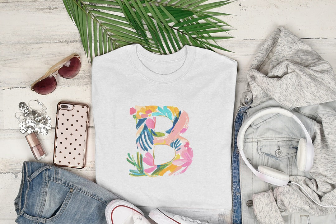 White t-shirt with a colorful floral letter B graphic, laid flat with clothing and accessories.