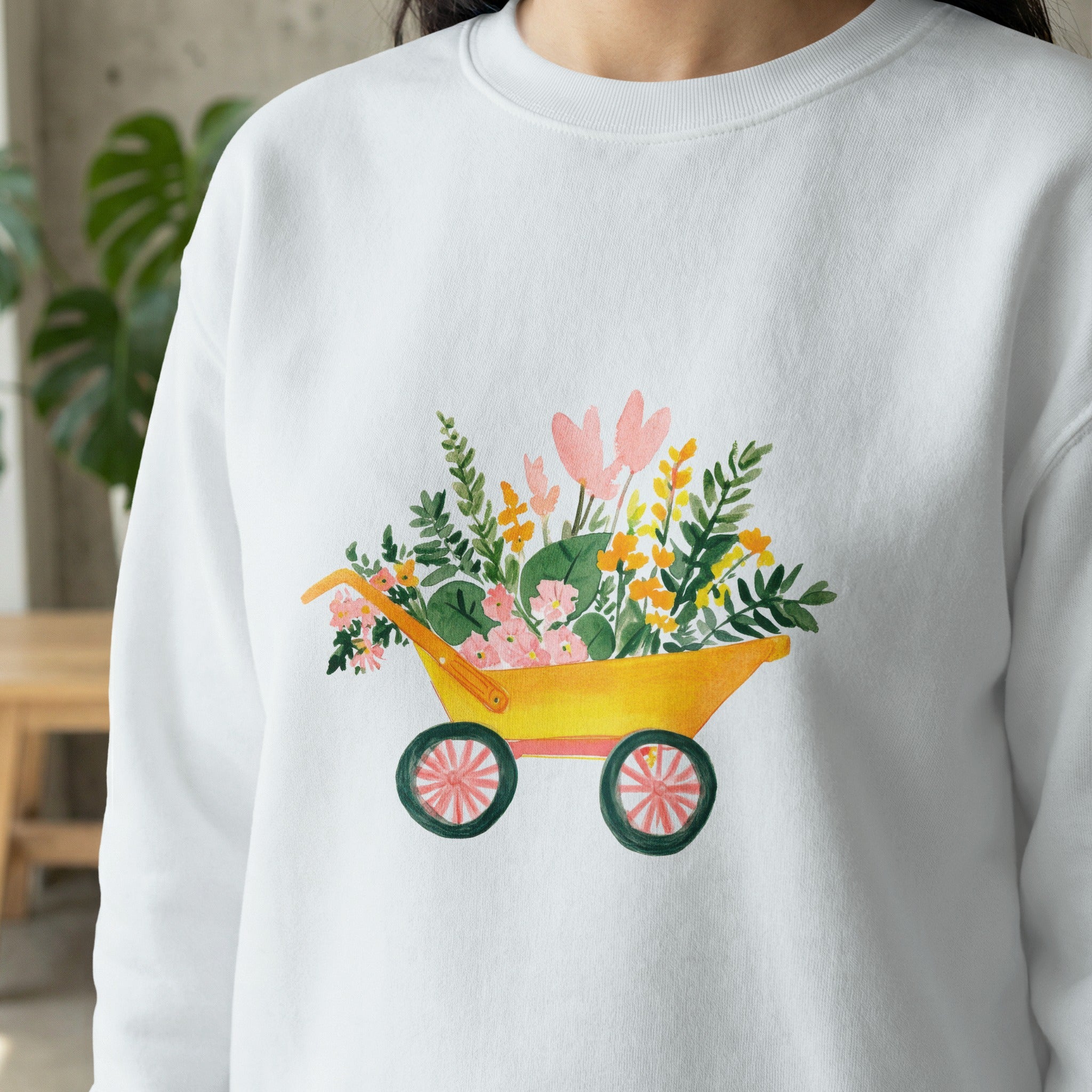 White sweatshirt with a watercolor illustration of a yellow wheelbarrow filled with flowers.