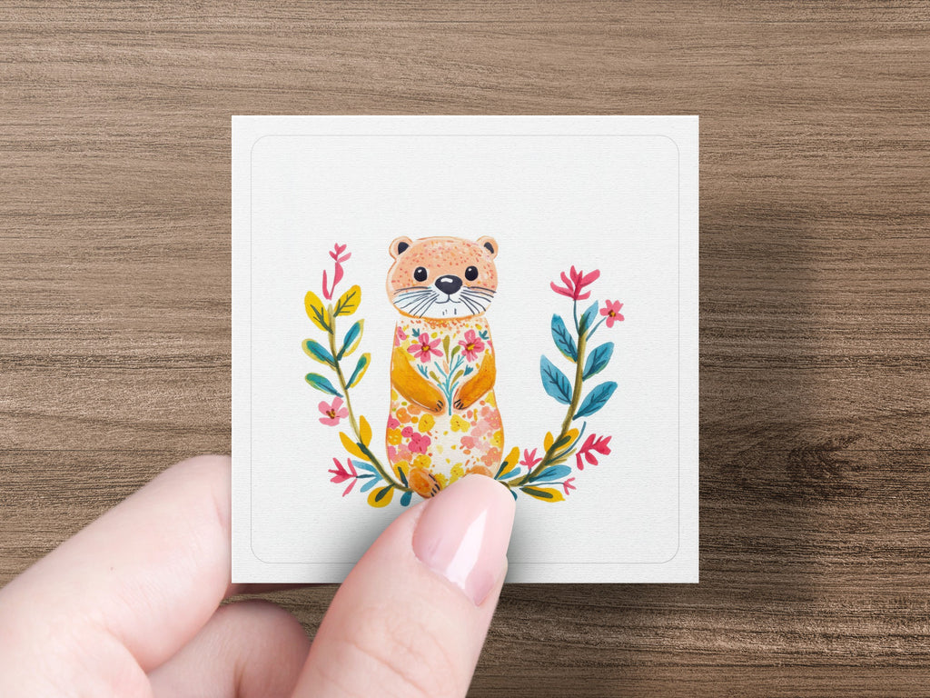 Watercolor otter illustration with floral wreath on square card held by a hand.