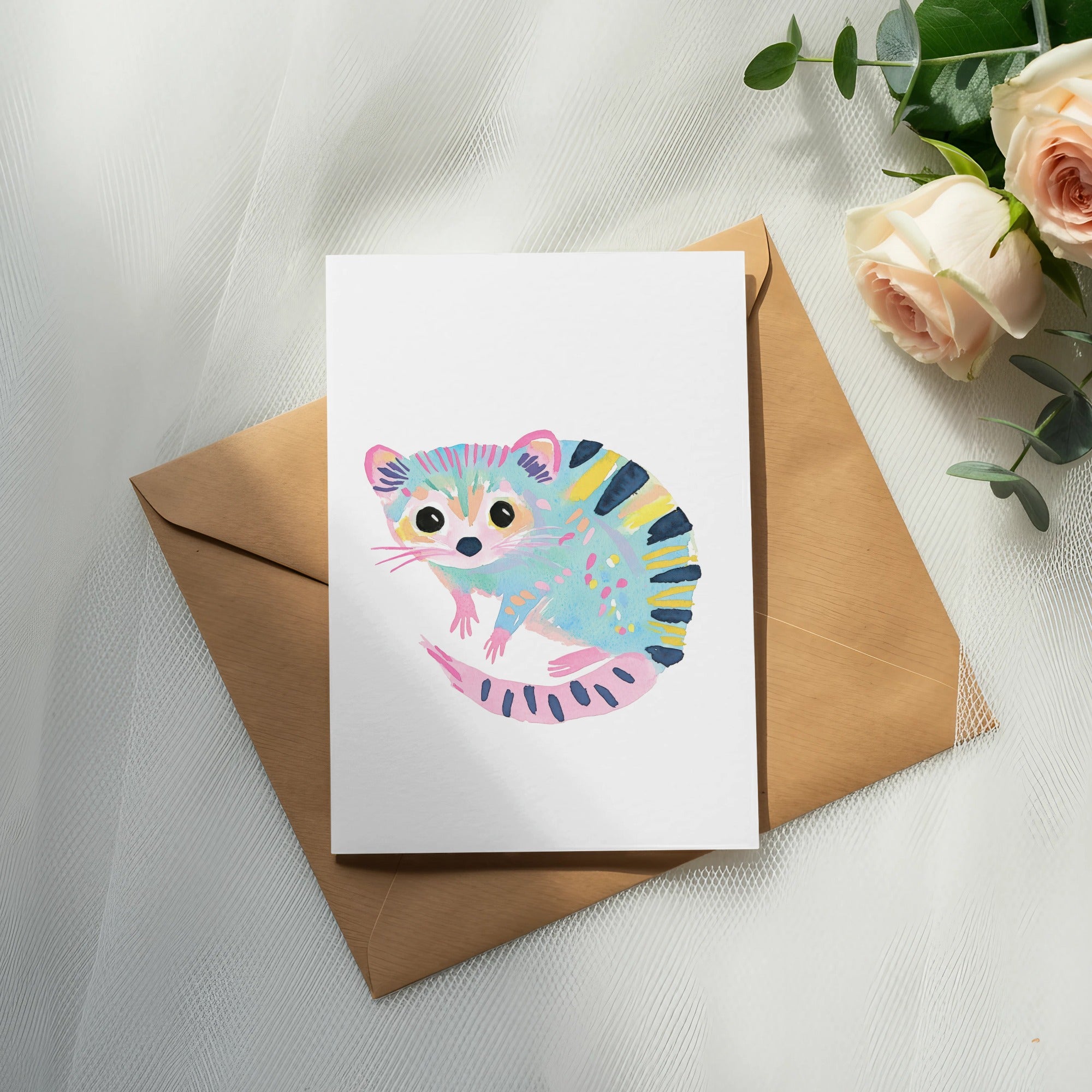 Card with watercolor raccoon illustration on brown envelope, with roses and eucalyptus.