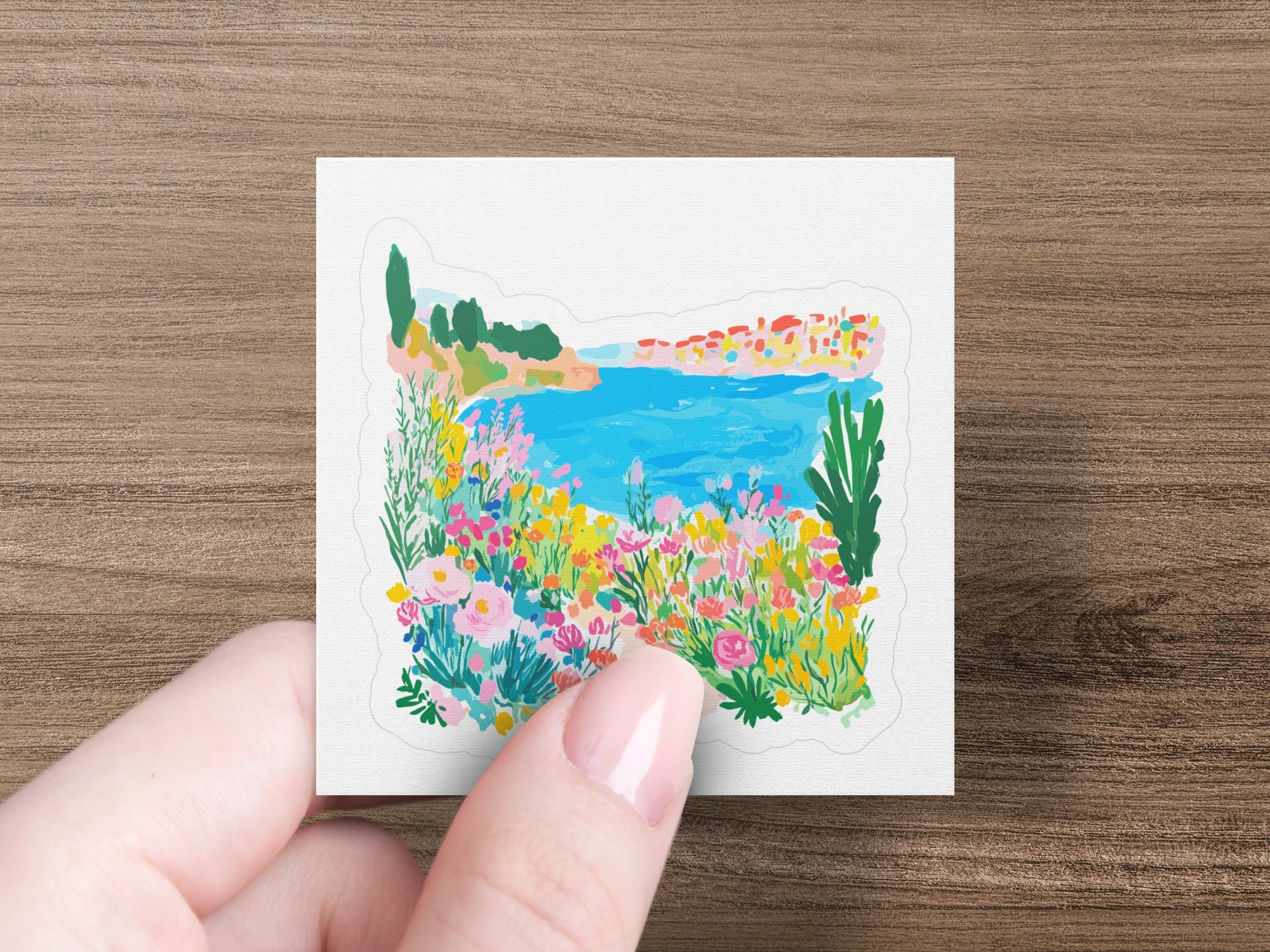 Hand holding a sticker depicting a colorful Mediterranean coastal village with flowers.