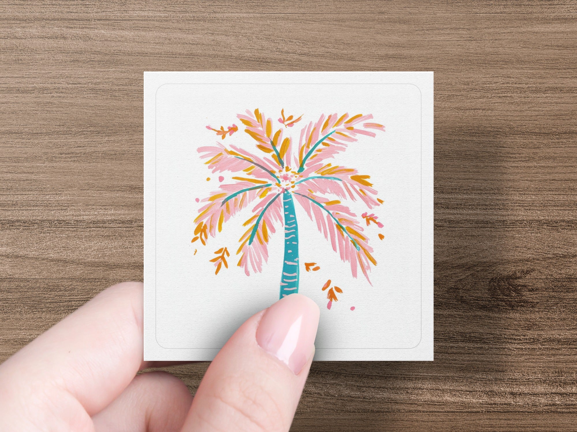 Pink and blue palm tree illustration on a white card held by a person's fingers.