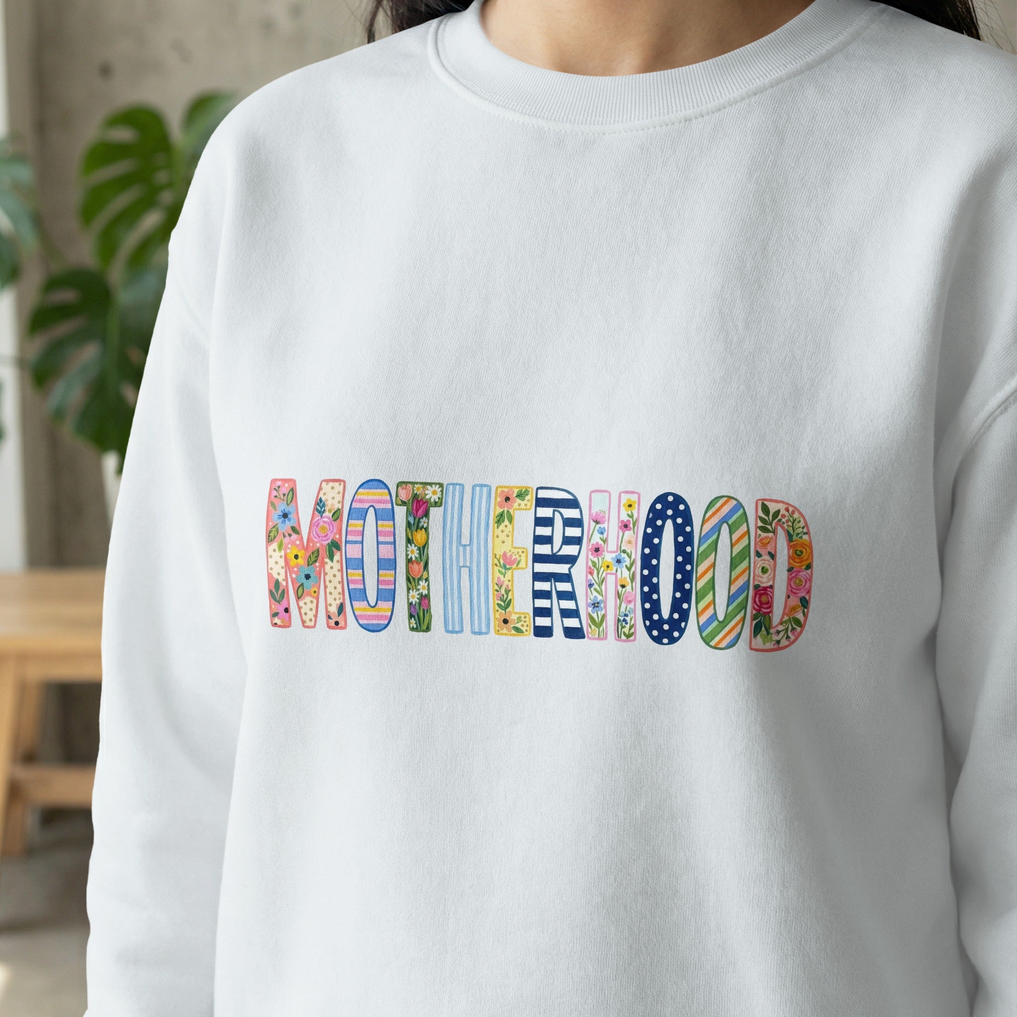 White sweatshirt with "MOTHERHOOD" in colorful, patterned block letters.