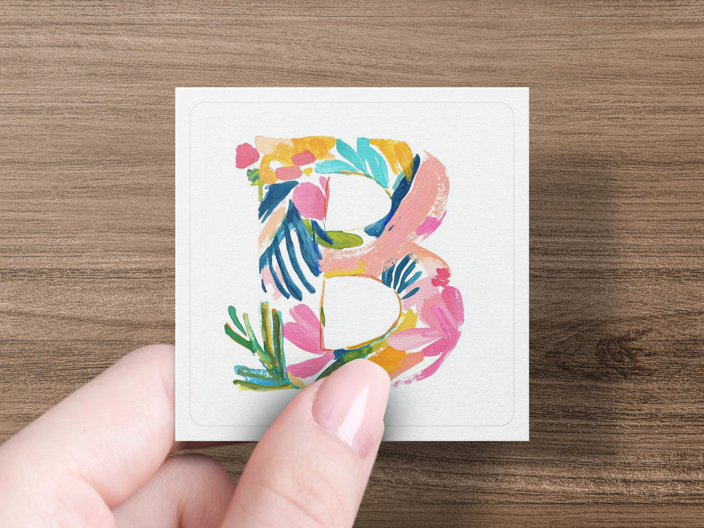 Hand holding a square card with a colorful floral "B" illustration.