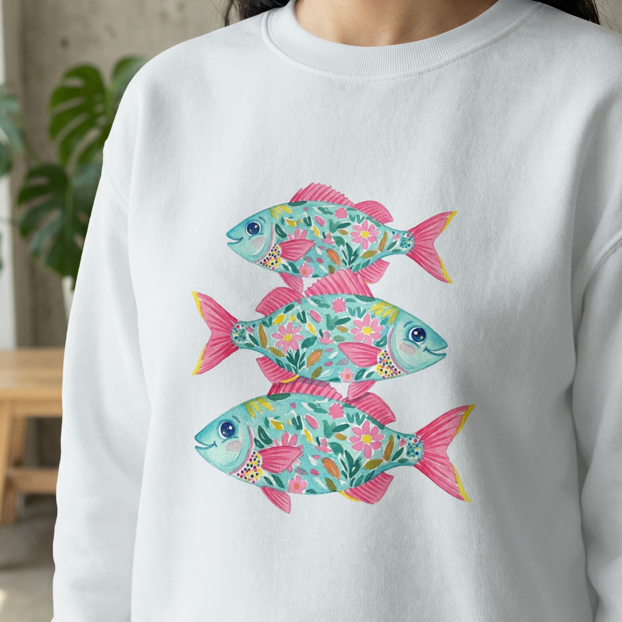 Three stylized teal fish with pink floral patterns stacked vertically on a white sweatshirt.