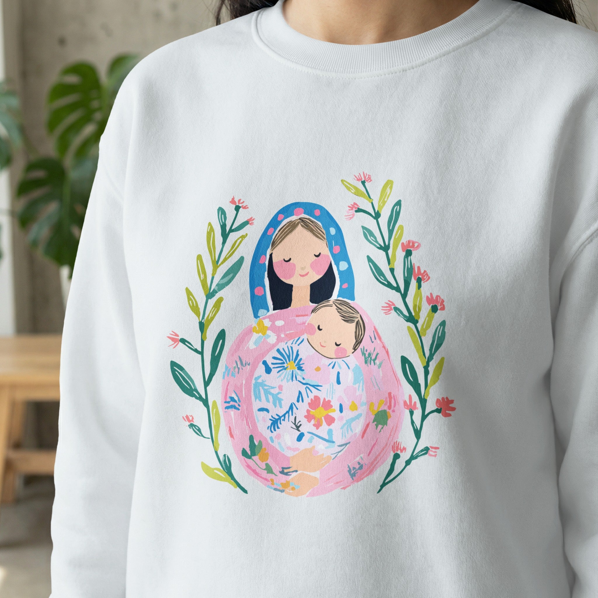 White sweatshirt with a colorful floral illustration of Mary holding baby Jesus.