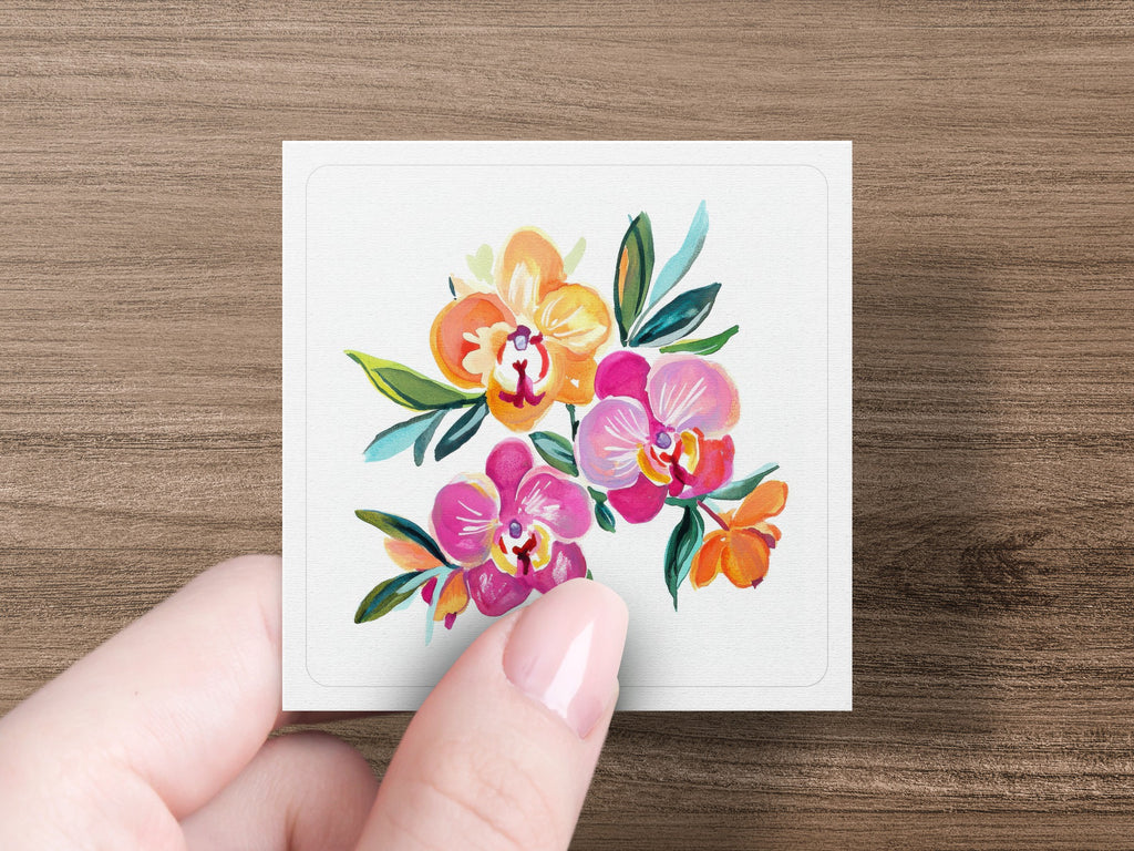 Hand holding a square card featuring a colorful watercolor painting of orchids.