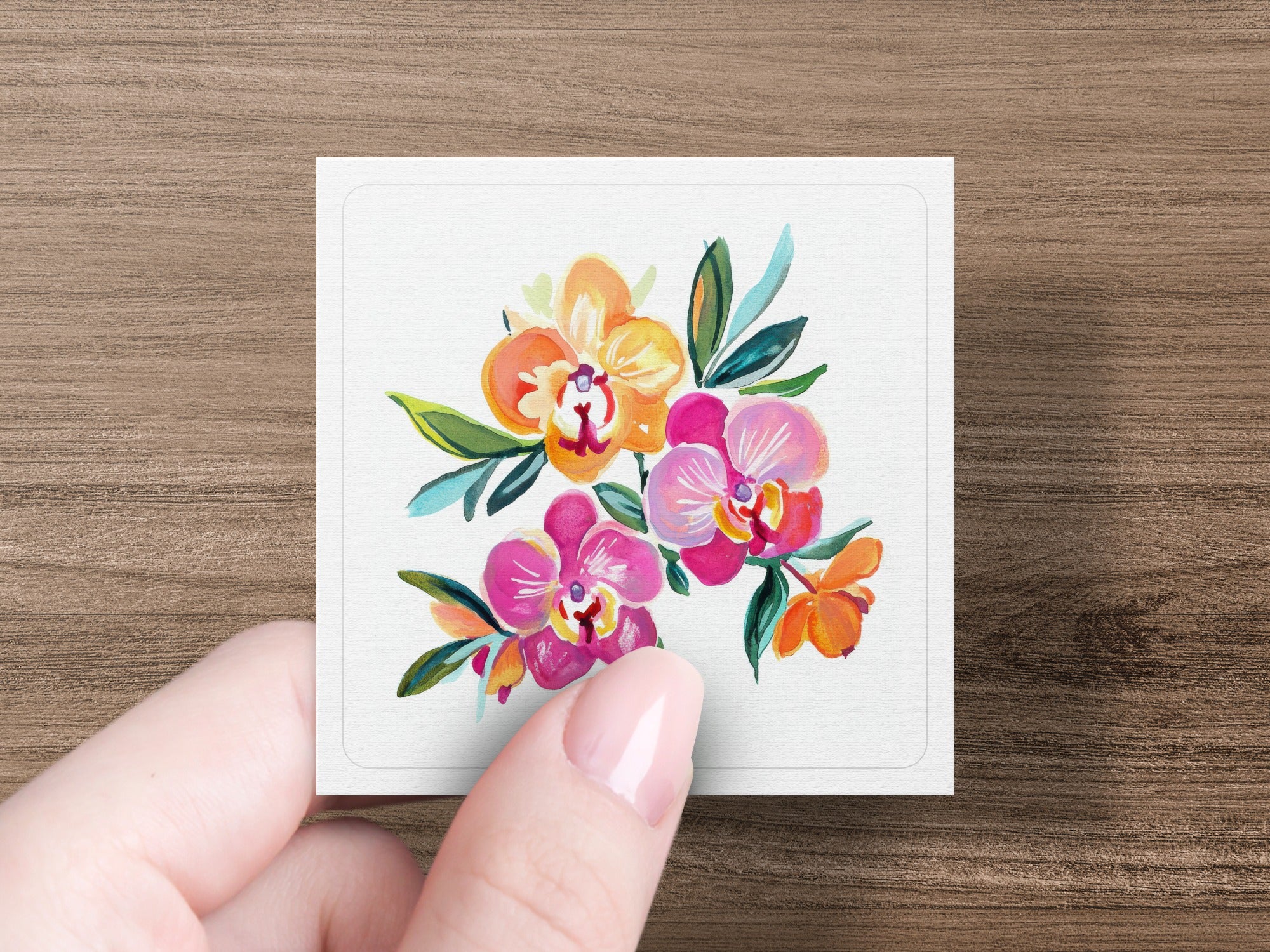 Hand holding a square card featuring a colorful watercolor painting of orchids.