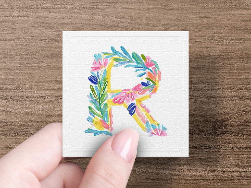 Hand holding a square card with a floral watercolor letter R.
