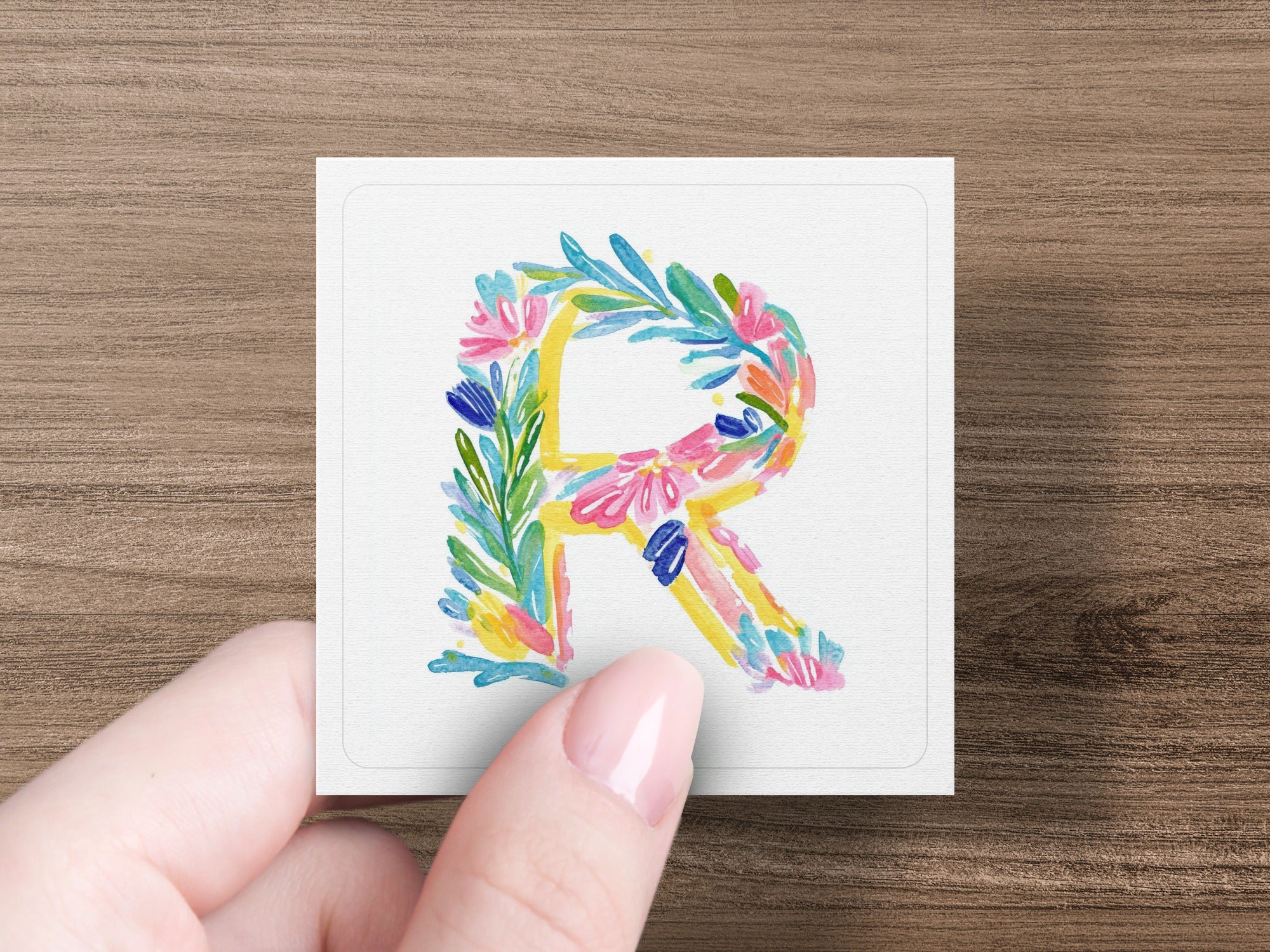 Hand holding a square card with a floral watercolor letter R.