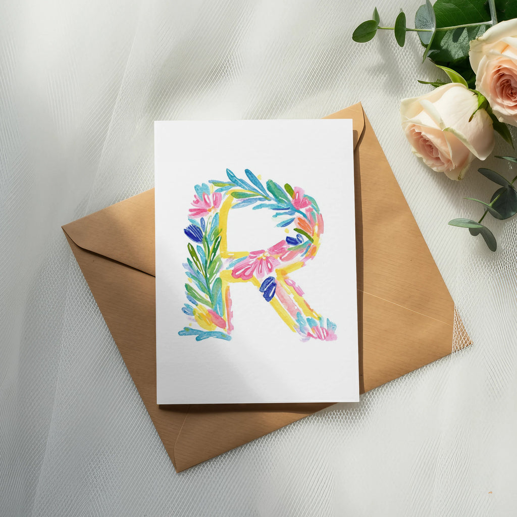 Watercolor letter R with floral accents on a card atop a brown envelope.