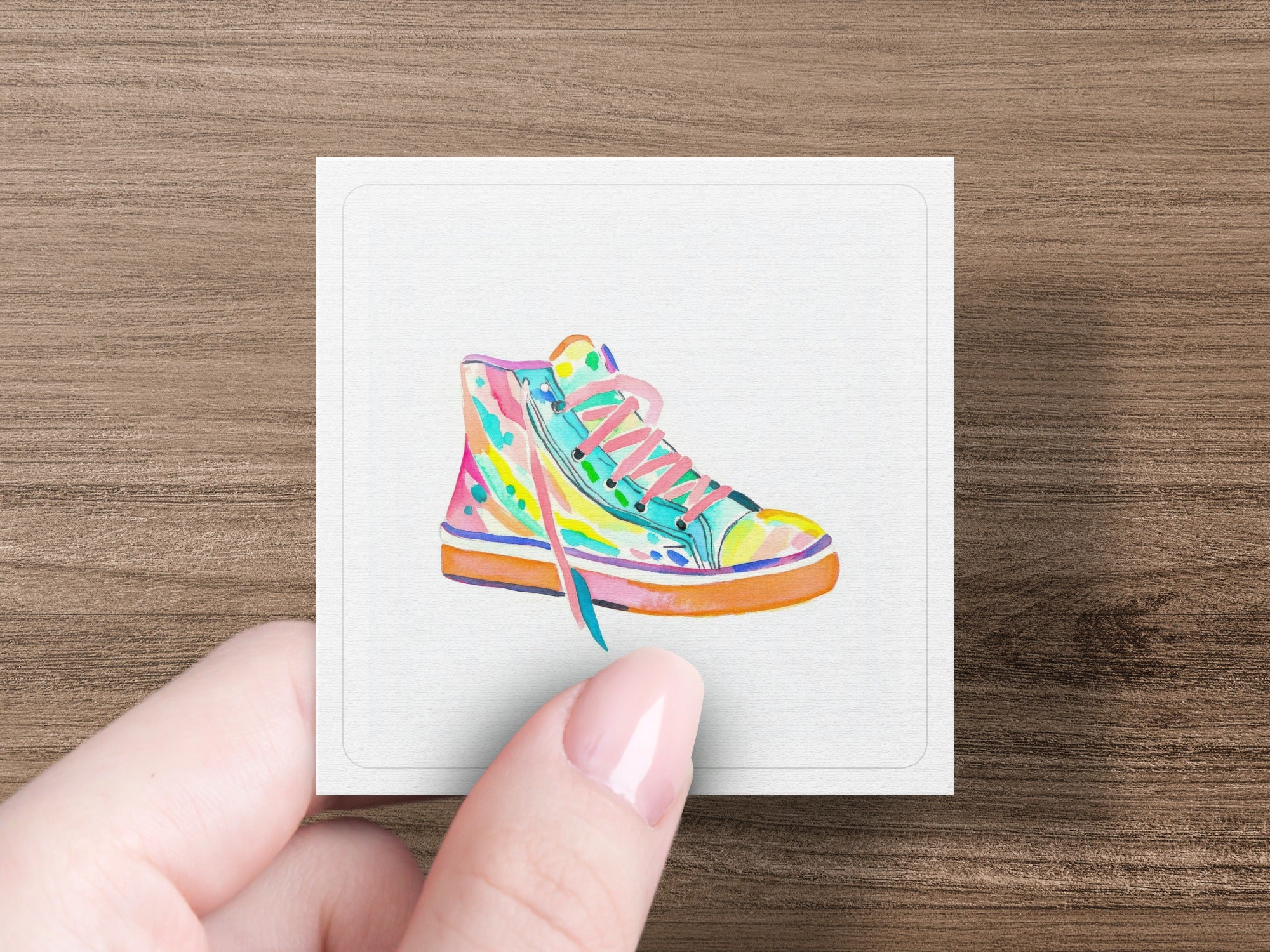 Colorful high-top sneaker illustration on a white card held by a hand.
