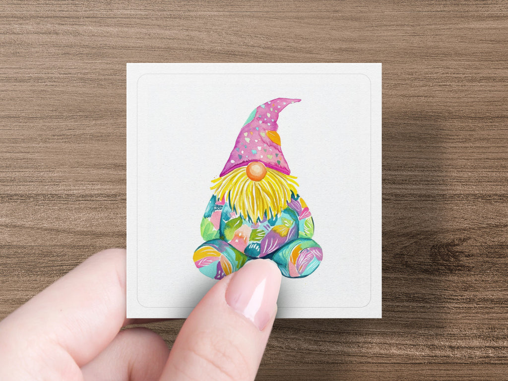 Watercolor gnome with colorful patterned hat and clothing, held by a hand.