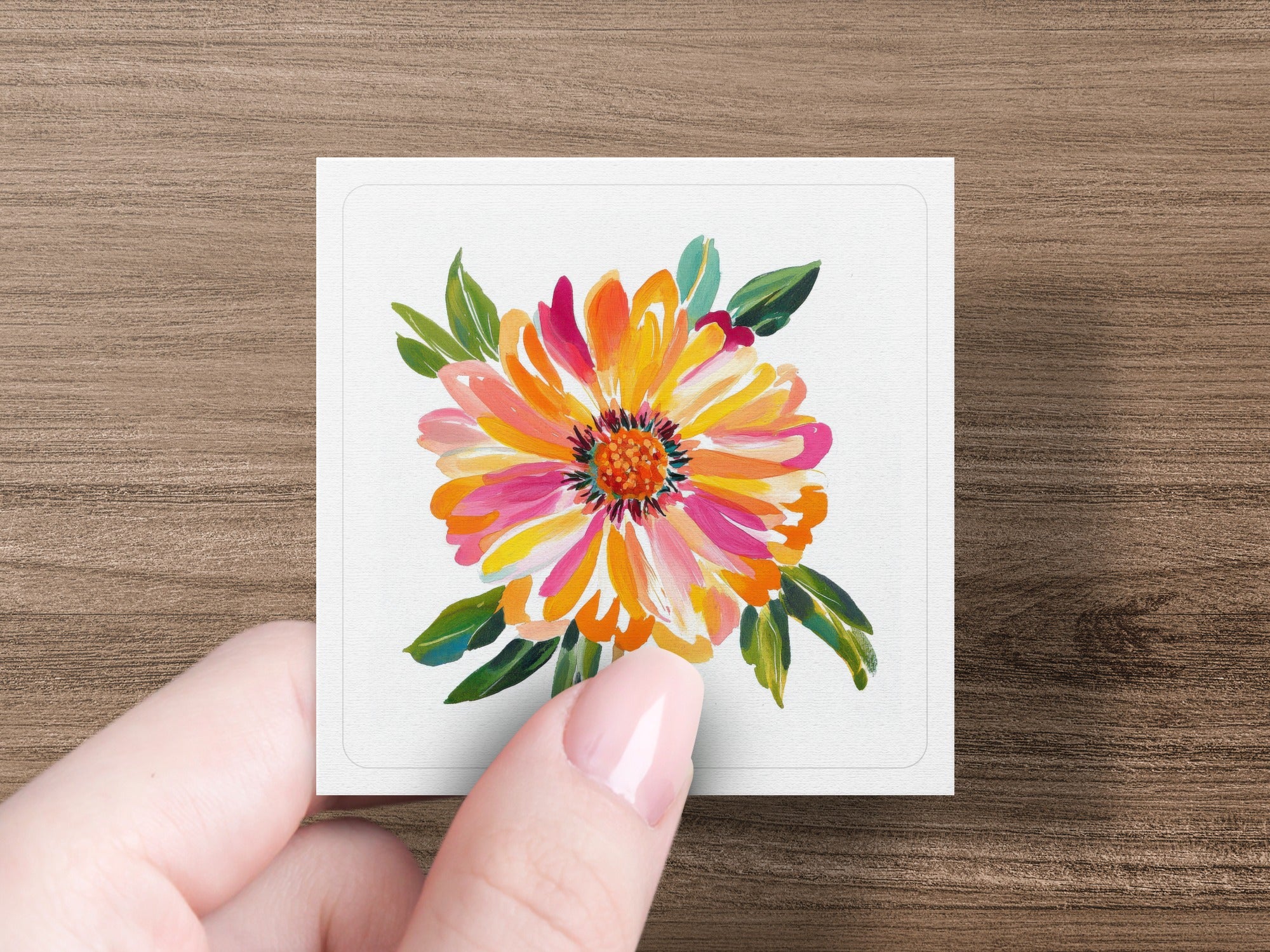 Hand holding square card with vibrant floral painting of a dahlia.