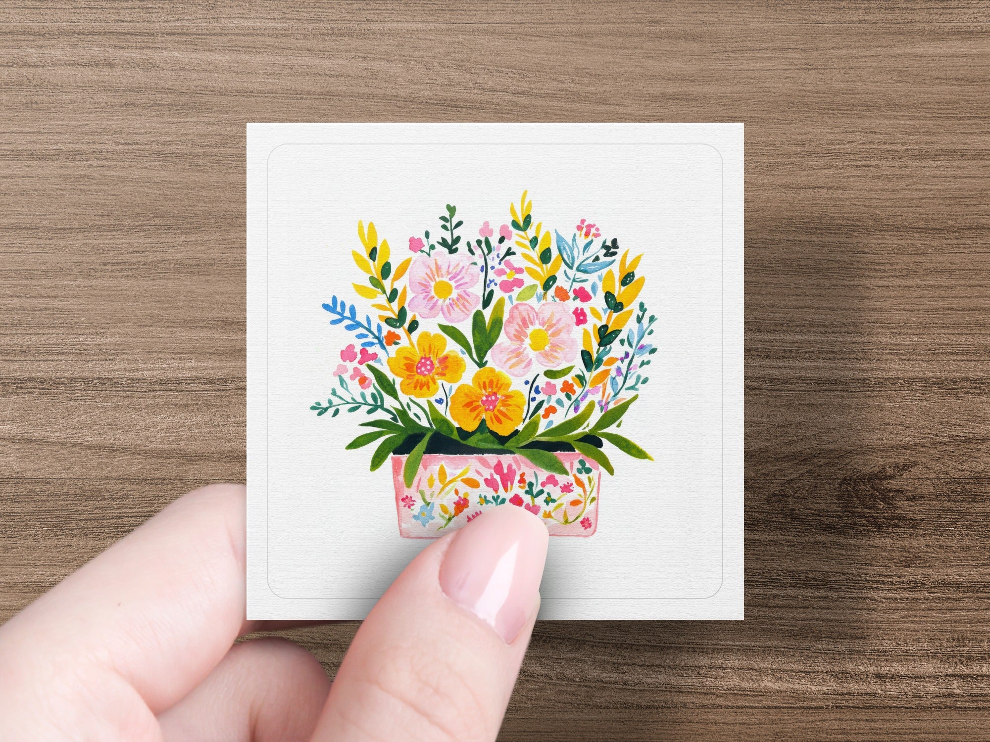 Pink floral pot with colorful blossoms and leaves, held by a hand.