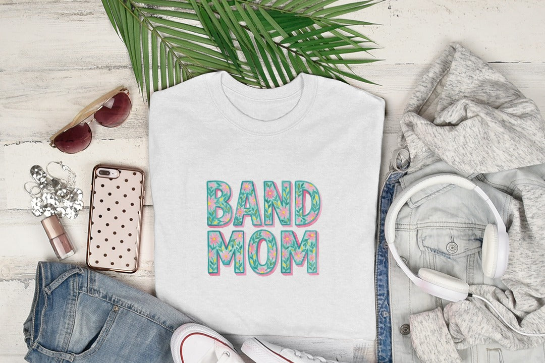 White t-shirt with "BAND MOM" in floral letters, displayed with jeans and headphones.