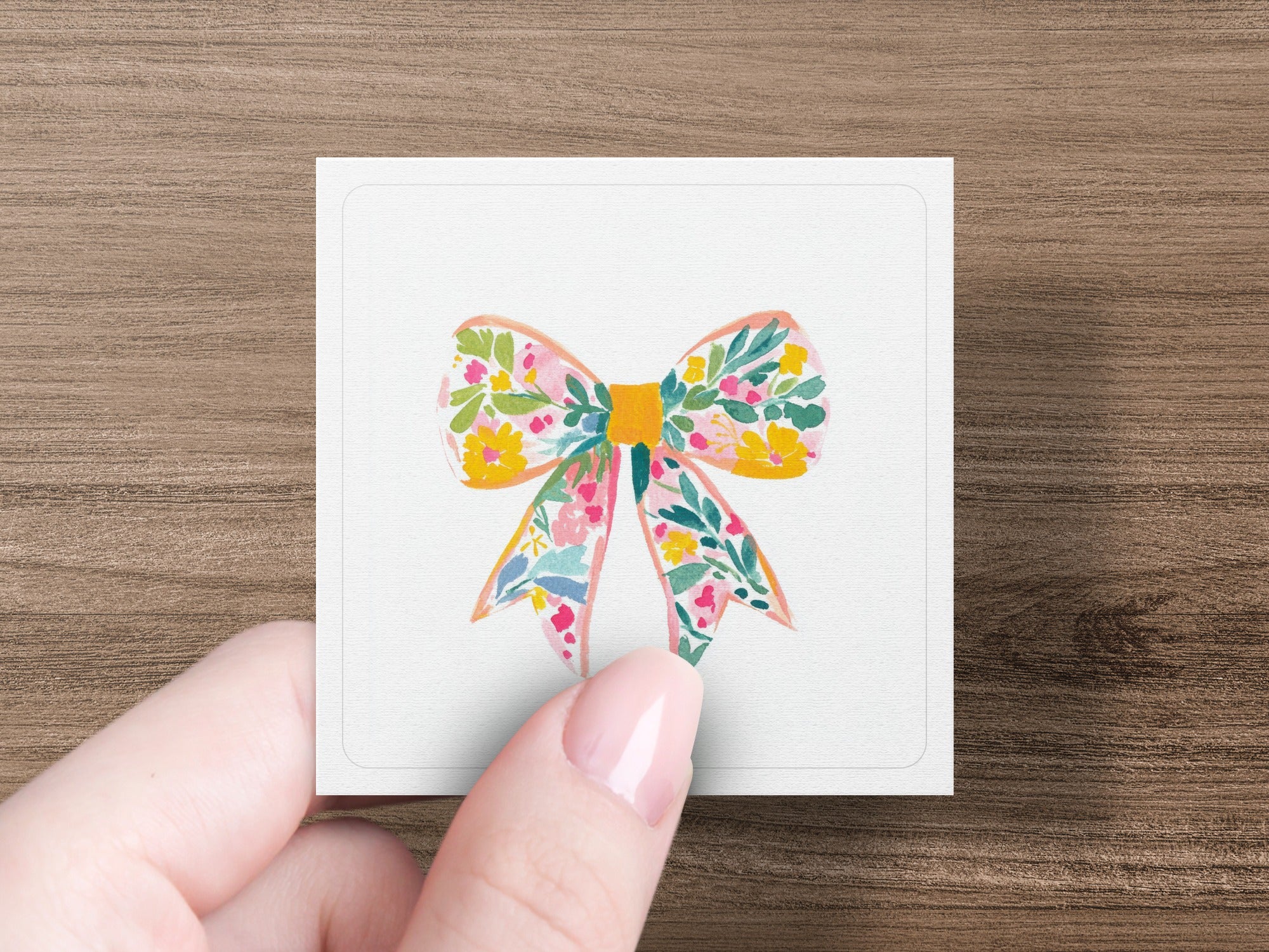 Floral bow illustration on a white square card held by a hand.