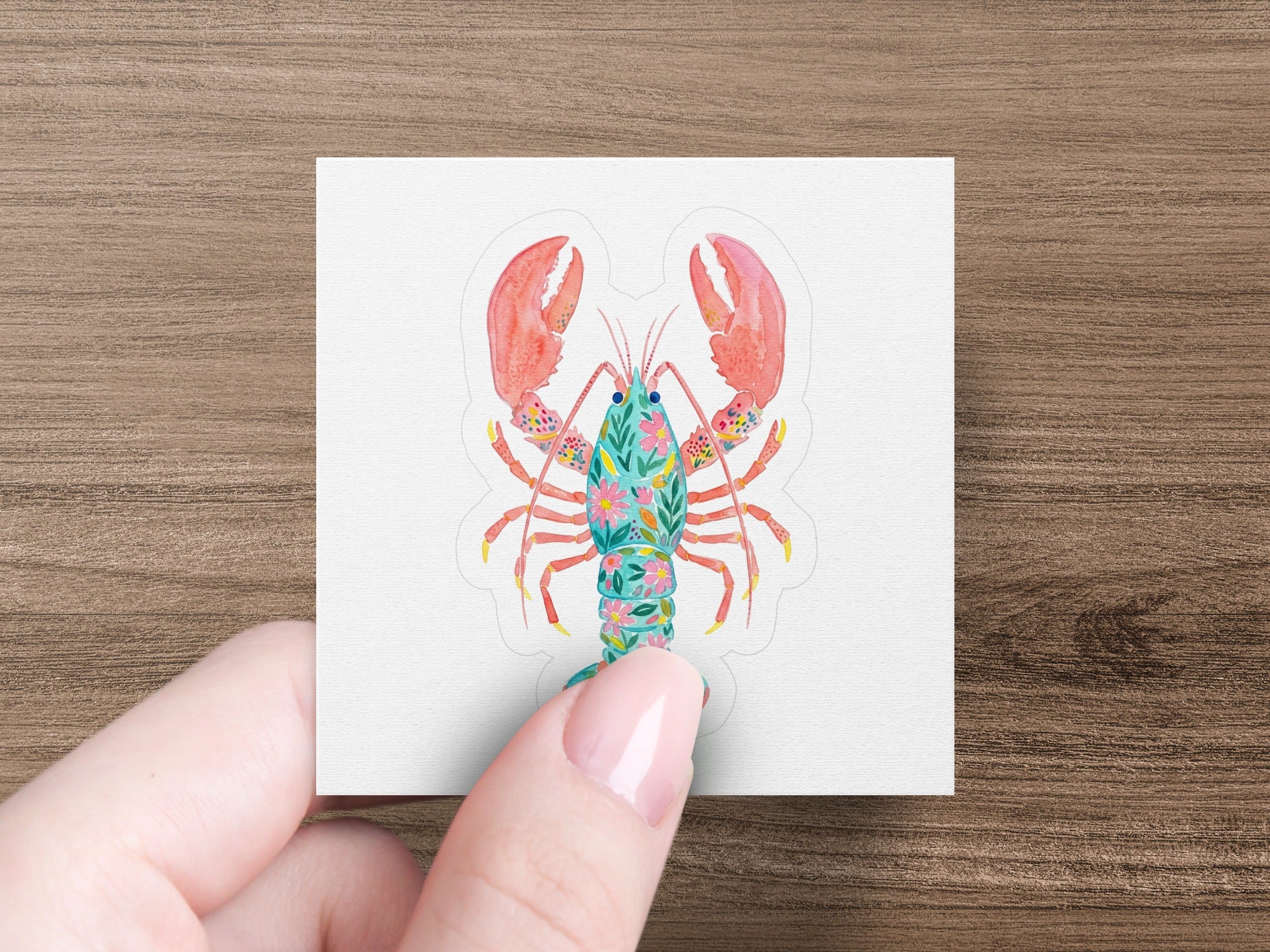 Pinky finger holds a square sticker of a floral patterned lobster.