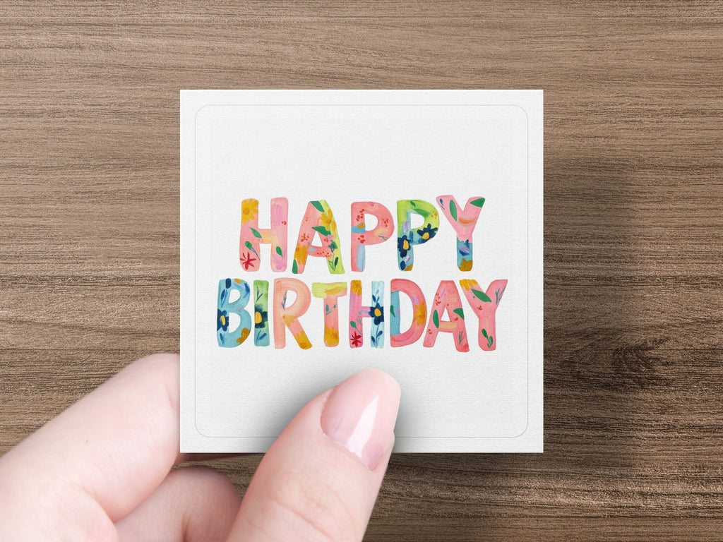 Hand holding square card with "Happy Birthday" in colorful floral lettering.