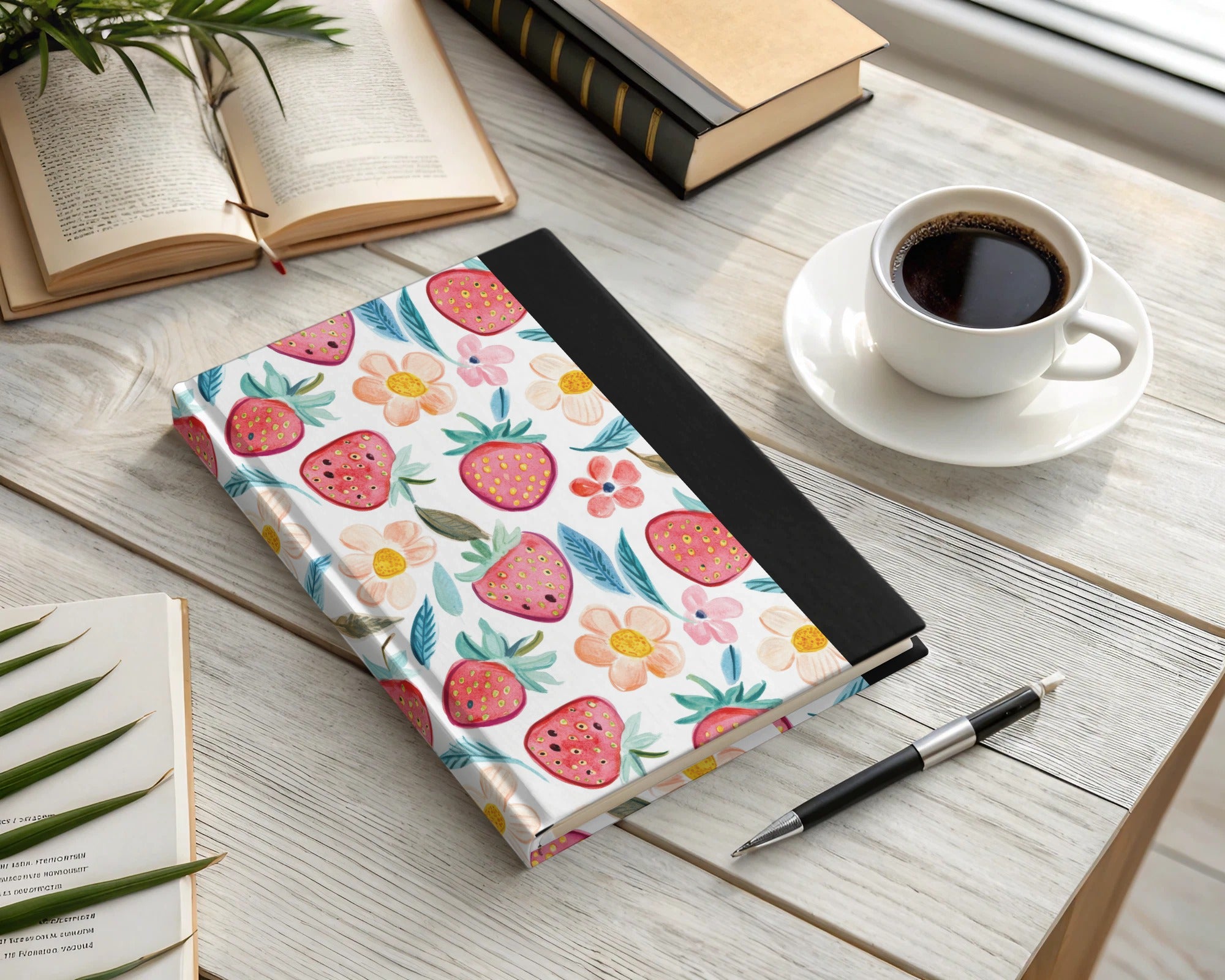 Journal with strawberry and floral pattern, pen, and coffee on a wooden desk.