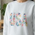White sweatshirt with "USA" in colorful floral watercolor letters.
