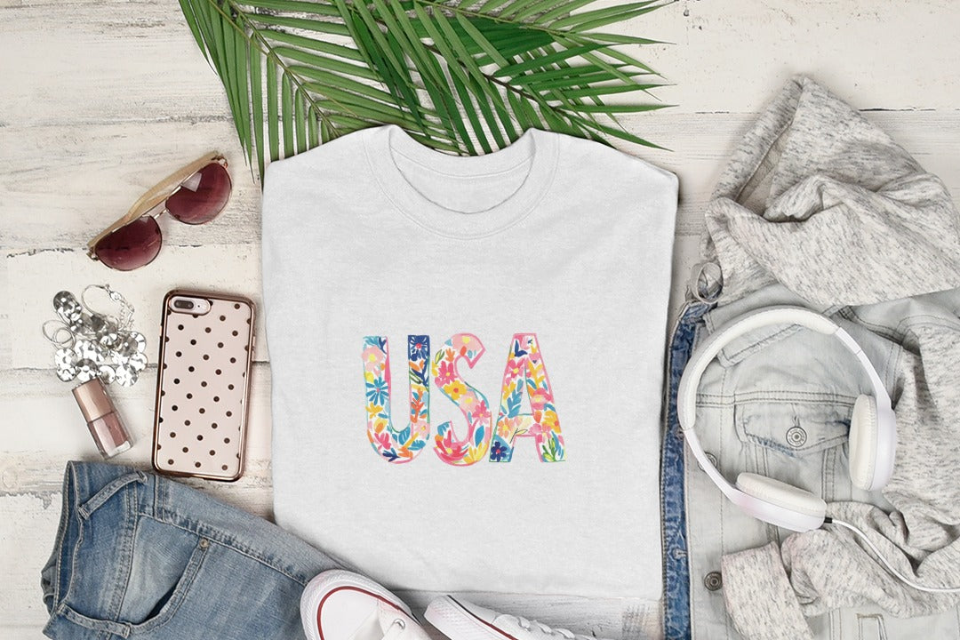 White t-shirt with colorful floral "USA" text, flat lay with headphones and sunglasses.