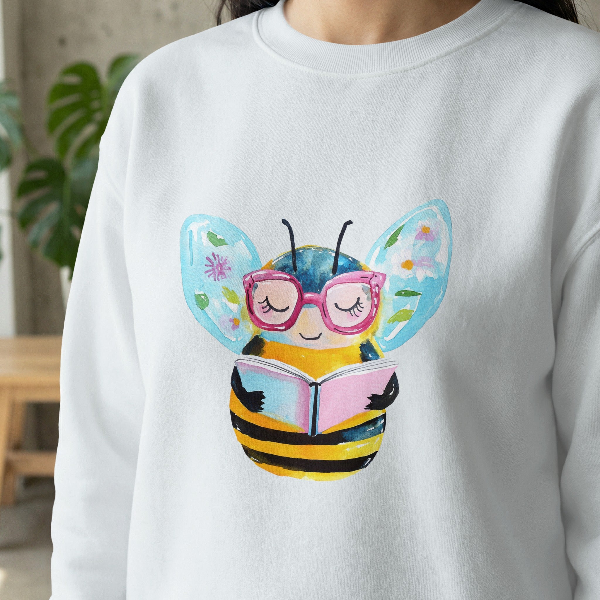 White sweatshirt with watercolor bee wearing glasses reading a book.