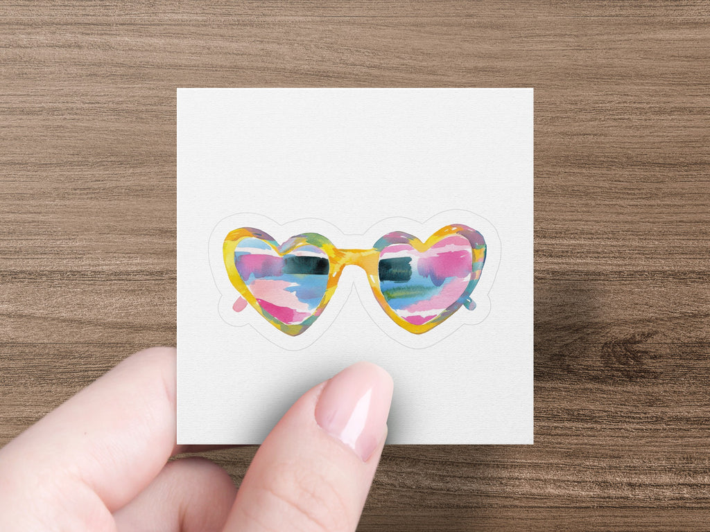 Heart-shaped sunglasses with colorful abstract pattern, held by a person's fingers.